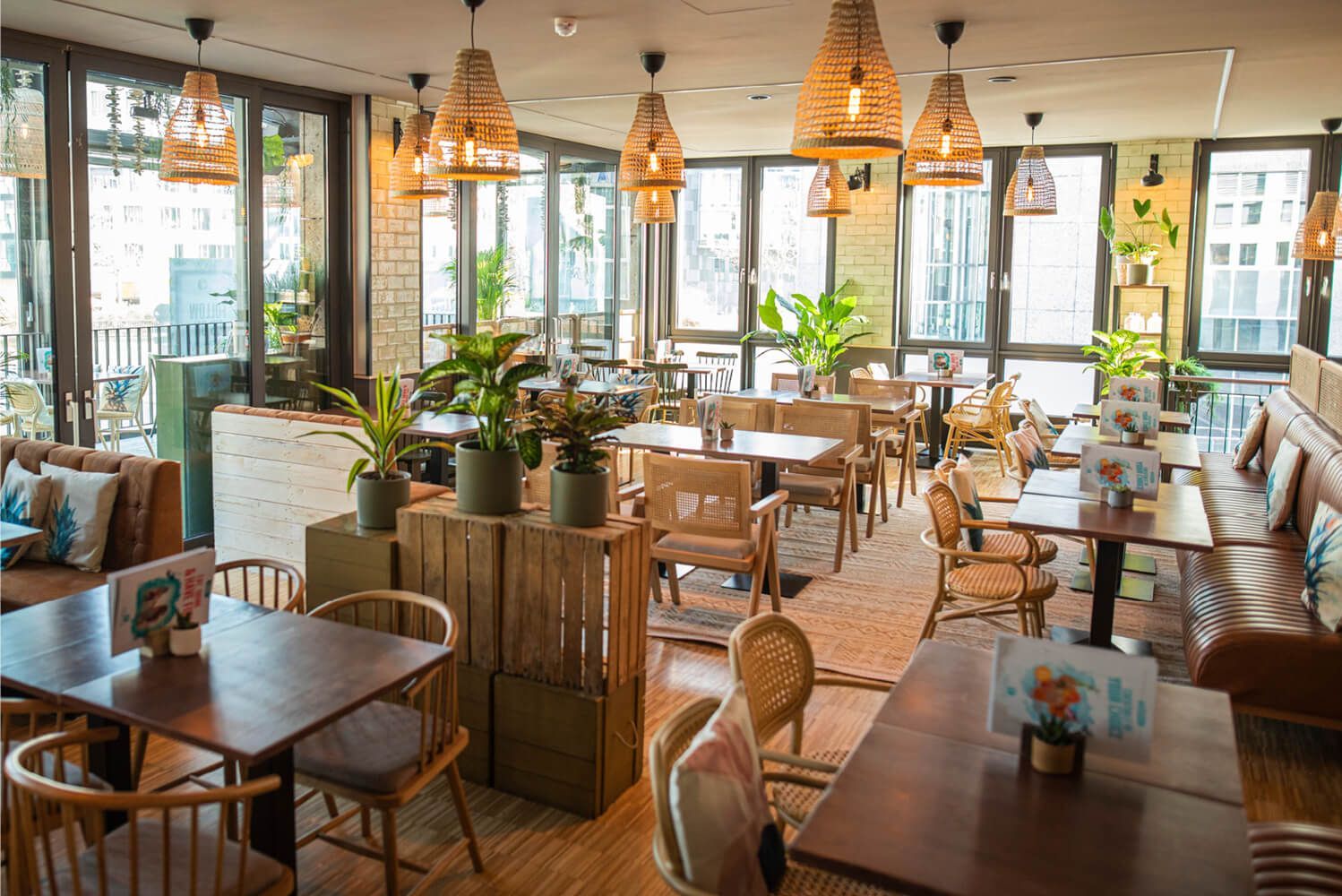 Helles, modernes Restaurant-Interieur mit Holztischen, Korbstühlen, Hängelampen, großen Fenstern, Grünpflanzen und natürlichem Licht. slide-3