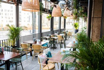 Modernes Straßencafé mit Holztischen, Rattanstühlen, farbenfrohen Pflanzen und großen Glasfenstern, die für eine helle, luftige Atmosphäre sorgen. slide-2